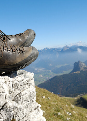 Das Bild zeigt eine Person, die auf einem Berg sitzt und nach unten blickt. Allerdings sind nur die ledernen Bergschuhe und die Wadern der Person zu sehen. Weiter unten erkennt man Bergwiesen, Wälder und ein Tal. Dahinter erheben sich weitere Berge. Der Himmel ist blau. Die Sonne scheint.  