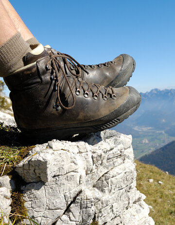 Das Bild zeigt eine Person, die auf einem Berg sitzt und nach unten blickt. Allerdings sind nur die ledernen Bergschuhe und die Wadern der Person zu sehen. Weiter unten erkennt man Bergwiesen, Wälder und ein Tal. Dahinter erheben sich weitere Berge. Der Himmel ist blau. Die Sonne scheint.  