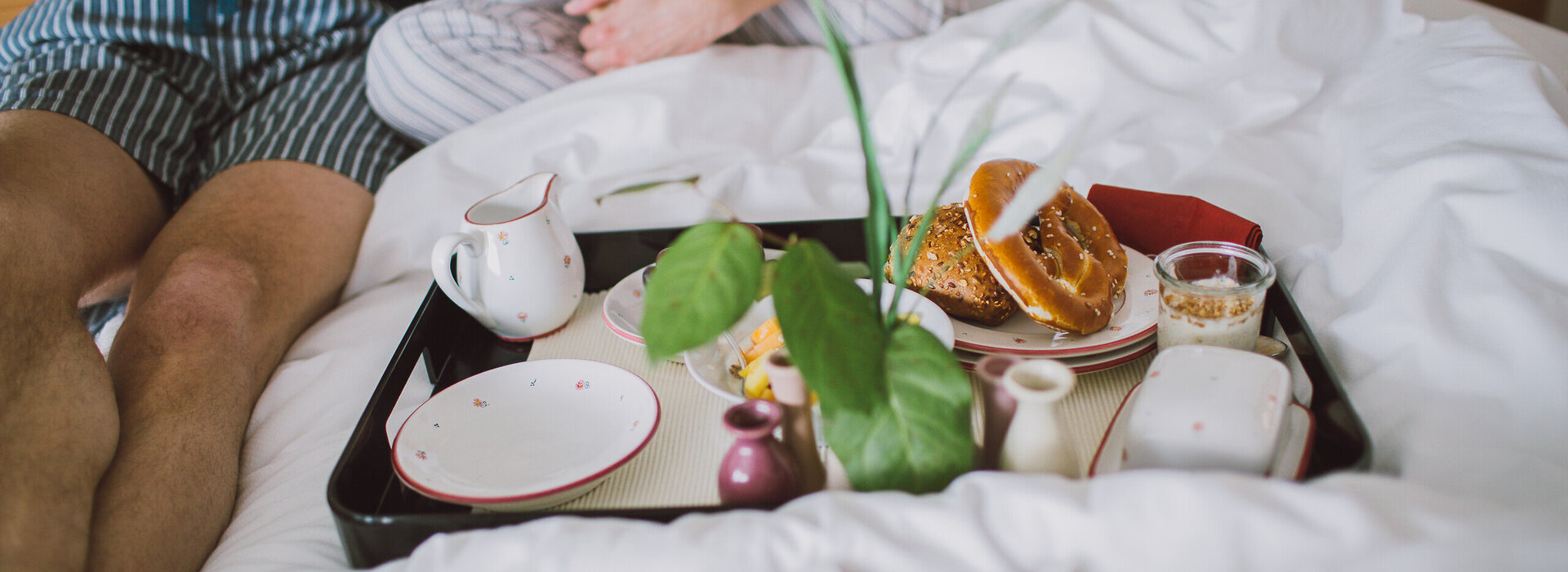 Das Bild zeigt ein Frühstückstablett, auf dem zwei Tellern, Tassen, Müsli, Butter, einem Kännchen stehen. Das Tablett steh auf einer weißen Bettdecke. Dahinter sitzen zwei Personen. Man kann nur die Beine der Personen sehen. Sie tragen Pyjamahosen.   
