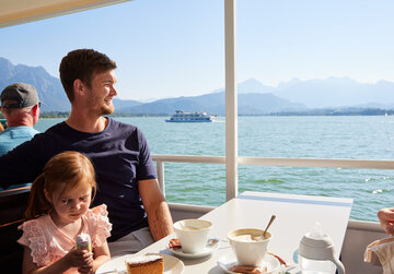 Das Bild fängt eine ruhige Szene auf einem Boot ein, wo ein Mann und ein junges Mädchen an einem Tisch sitzen und ein Essen gemeinsam genießen. Der Mann, der ein blaues Hemd trägt, sitzt auf der linken Seite des Tisches, während das Mädchen, das ein rosa Hemd trägt, auf der rechten Seite sitzt. Der Tisch ist mit einer weißen Tasse, einer weißen Schüssel und einer weißen Flasche geschmückt, was auf eine legere und entspannte Essenszeit hinweist.  Das Boot, auf dem sie sind, segelt über einem weiten Gewässer, mit majestätischen Bergen, die eine malerische Kulisse bilden. Der klare blaue Himmel über ihnen und das ruhige Wasser unter ihnen tragen zur Ruhe der Szene bei. Die Gesamtatmosphäre des Bildes ist eine von Frieden und Zusammengehörigkeit, wenn der Mann und das Mädchen ein Essen teilen und dabei die atemberaubende Umgebung um sie herum genießen.