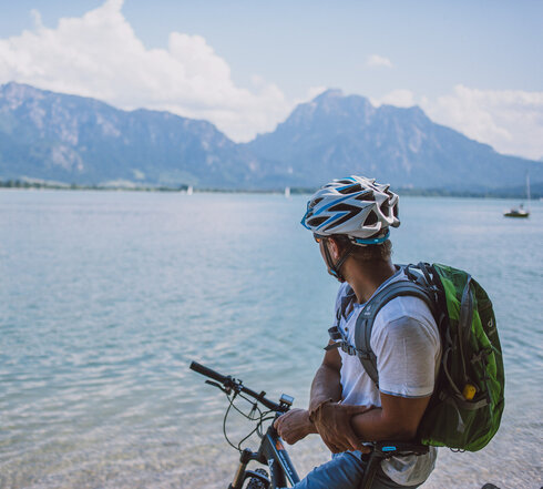 Das Bild fängt einen ruhigen Moment eines Radfahrers ein, der eine Pause von seiner Reise macht. Der Radfahrer lehnt auf seinem Fahrrad, das auf einem sandigen Strand geparkt ist. Der Radfahrer blickt auf den vor ihm liegenden Forggensee. Das Wasser ist ein tiefes Blau, das den klaren blauen Himmel darüber reflektiert. In der Ferne erheben sich majestätische Berge, die der Szene ein Gefühl von Größe verleihen. Der Radfahrer trägt einen Helm. Die Bild vermittelt eine ruhige Stimmung.