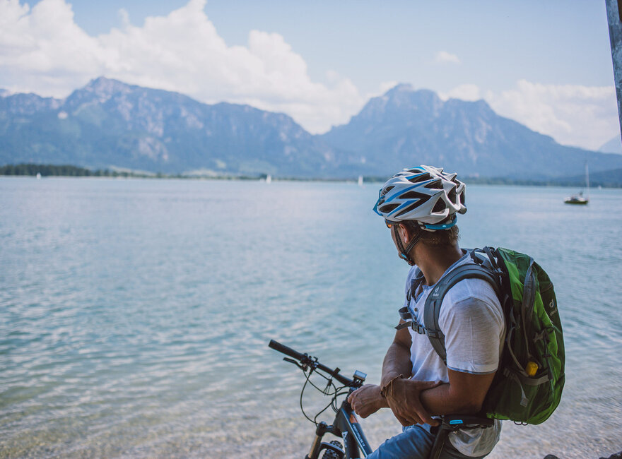 Das Bild fängt einen ruhigen Moment eines Radfahrers ein, der eine Pause von seiner Reise macht. Der Radfahrer lehnt auf seinem Fahrrad, das auf einem sandigen Strand geparkt ist. Der Radfahrer blickt auf den vor ihm liegenden Forggensee. Das Wasser ist ein tiefes Blau, das den klaren blauen Himmel darüber reflektiert. In der Ferne erheben sich majestätische Berge, die der Szene ein Gefühl von Größe verleihen. Der Radfahrer trägt einen Helm. Die Bild vermittelt eine ruhige Stimmung.