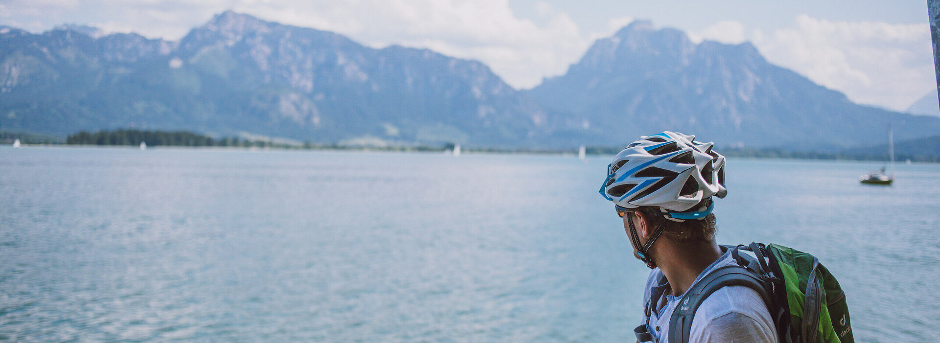 Das Bild fängt einen ruhigen Moment eines Radfahrers ein, der eine Pause von seiner Reise macht. Der Radfahrer lehnt auf seinem Fahrrad, das auf einem sandigen Strand geparkt ist. Der Radfahrer blickt auf den vor ihm liegenden Forggensee. Das Wasser ist ein tiefes Blau, das den klaren blauen Himmel darüber reflektiert. In der Ferne erheben sich majestätische Berge, die der Szene ein Gefühl von Größe verleihen. Der Radfahrer trägt einen Helm. Die Bild vermittelt eine ruhige Stimmung.