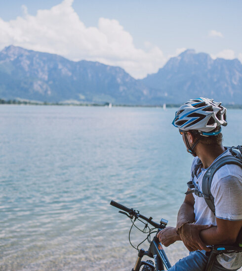 Das Bild fängt einen ruhigen Moment eines Radfahrers ein, der eine Pause von seiner Reise macht. Der Radfahrer lehnt auf seinem Fahrrad, das auf einem sandigen Strand geparkt ist. Der Radfahrer blickt auf den vor ihm liegenden Forggensee. Das Wasser ist ein tiefes Blau, das den klaren blauen Himmel darüber reflektiert. In der Ferne erheben sich majestätische Berge, die der Szene ein Gefühl von Größe verleihen. Der Radfahrer trägt einen Helm. Die Bild vermittelt eine ruhige Stimmung.
