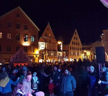 Das Bild zeigt viele Menschen auf einem Platz. Im Hintergrund sieht man beleuchtete Häuserfassaden. Die Menschen unterhalten sich. Alle tragen warme Winterklamotten. Im Vordergrund kann man eine beleuchtete Bühne erahnen. Auf dem Platz stehen mehrere Weihnachtsmarktbuden. Es ist Abend.  