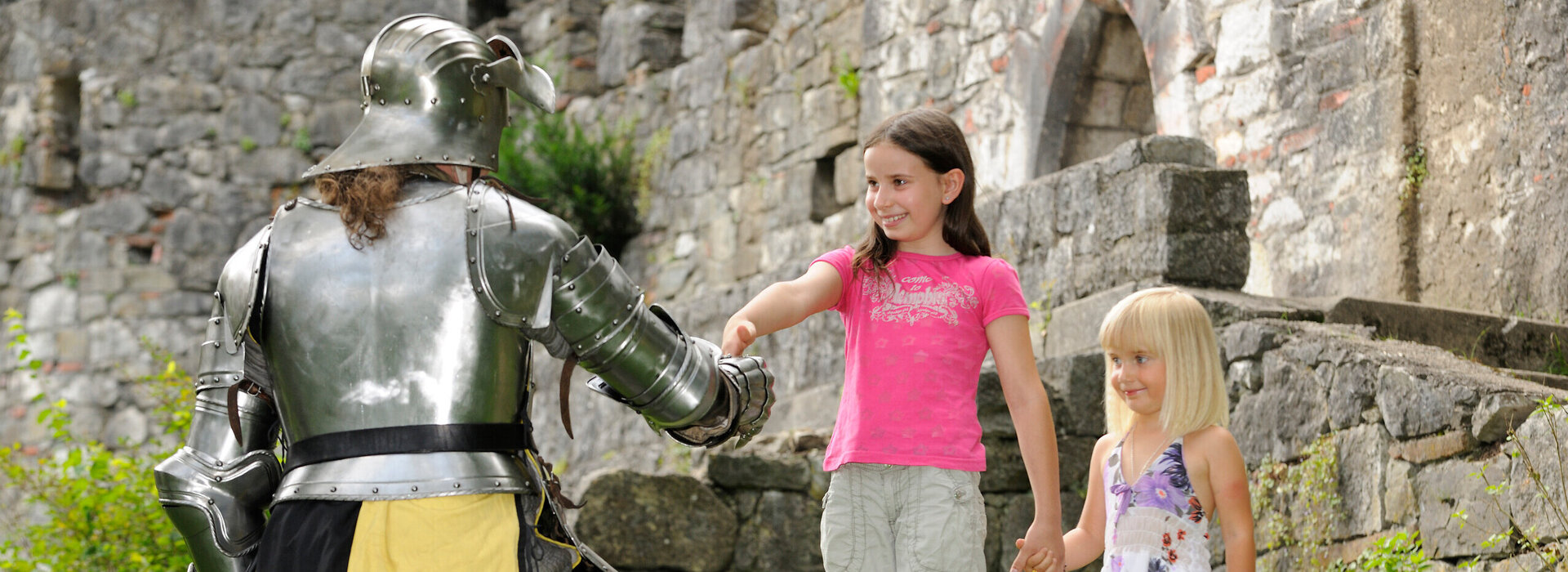 Das Bild zeigt einen Moment der Interaktion zwischen einem Ritter und zwei jungen Mädchen. Der Ritter in voller Rüstung kniet auf einem Knie in einer grünen Fläche. Er streckt seine Hand den Mädchen entgegen, die vor ihm stehen. Die Mädchen, in legerer Kleidung, halten Hände und scheinen mit dem Ritter zu sprechen. Die Kulisse des Bildes ist eine Steinmauer, die dem Bild einen historischen Kontext verleiht. Die Ritterrüstung und die Steinmauer lassen darauf schließen, dass die Szene in einem historischen oder mittelalterlichen Rahmen angesiedelt sein könnte. Die legere Kleidung der Mädchen kontrastiert mit der Rüstung des Ritters und hebt den Unterschied ihrer Rollen und der Zeitperioden, die sie repräsentieren, hervor. Das Bild enthält keine Texte.