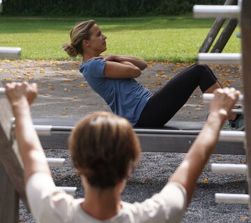 Man blickt auf zwei Frauen, die Outdoorsportgeräte nutzen. Sie tragen Sportkleidung. Eine Frau macht Situps und hat die Arme vor ihrem Oberkörper verschränkt. Die Sonne scheint.  