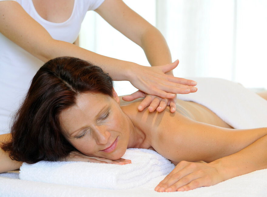 Das Bild zeigt eine Frau, die bäuchlings auf einer Massageliege liegt. Sie ist unbekleidet. Ein weißes Handtuch bedeckt ihr Gesäß und ihren unteren Rücken. Sie hat die Augen geschlossen. Über ihr steht eine Person, die ihr die obere Rückenpartie massiert. Die Person trägt ein weißes Oberteil. Der Raum ist hell erleuchtet.      