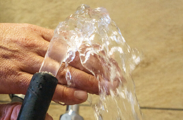 Das Bild zeigt zwei Hände die einen Wasserschlauch halten. Aus dem Wasserschlauch kommt ein klares Wasser, das über die eine Hand läuft. Im Hintergrund ist ein silberner Wasserhahn zu sehen.      