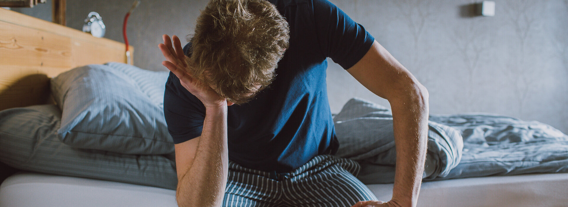 Das Bild zeigt einen Mann in einem kurzen Pyjama. Er sitzt am Bettrand hat die eine Hand auf den anderen Ellenbogen auf seine Knie gestützt. Mit der einen anderen Hand reibt er sich das Gesicht. 