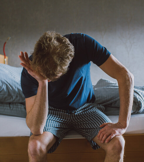 Das Bild zeigt einen Mann in einem kurzen Pyjama. Er sitzt am Bettrand hat die eine Hand auf den anderen Ellenbogen auf seine Knie gestützt. Mit der einen anderen Hand reibt er sich das Gesicht. 