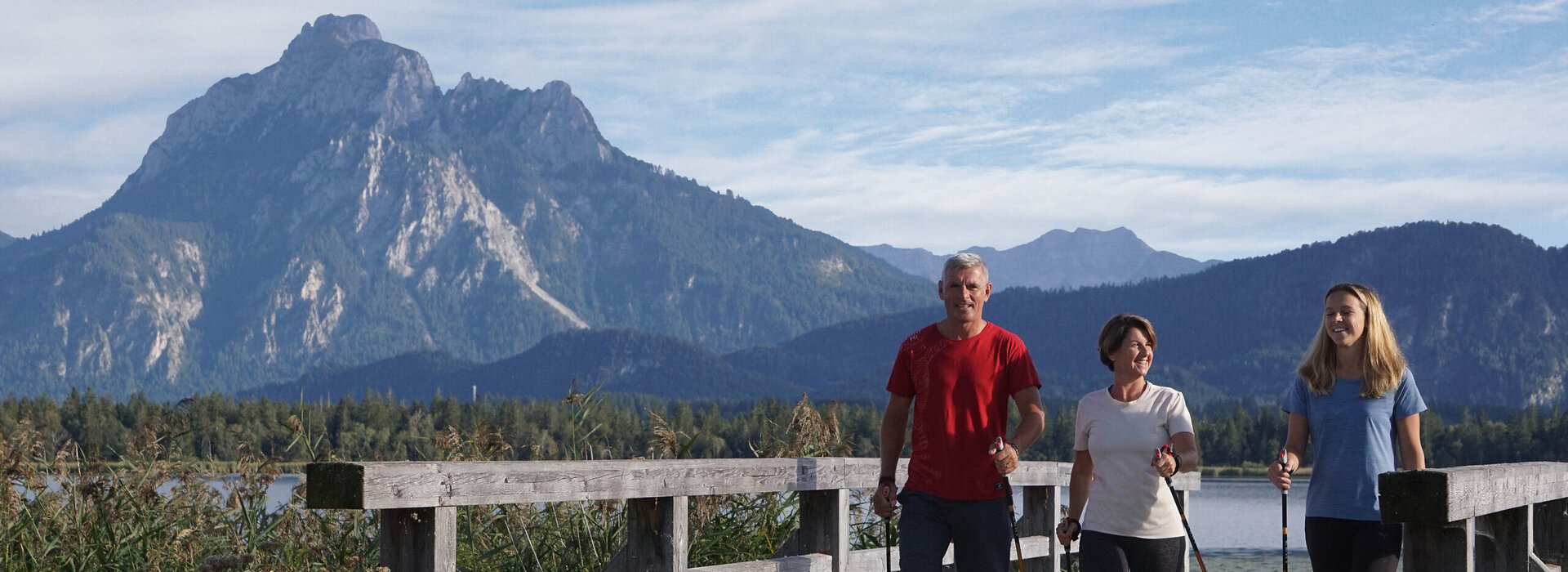 Das Bild zeigt drei Personen in Sportkleidung, die auf einer Holzbrücke laufen. Alle haben Nordic Walking Stöcke in den Händen. Sie lächeln. Im Hintergrund erheben sich hohe Berge und bewaldete Hügel.     