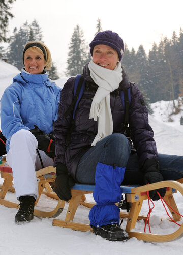Das Bild zeigt drei Frauen, die je auf einem Holzschlitten sitzen. Die Schlitten stehen hintereinander auf einer Piste. Alle tragen Winterkleidung und Mützen. Rundherum sind hohe Bäume zu sehen. Alle lächeln. Die Landschaft ringsum ist tief verschneit. Die Sonne scheint.