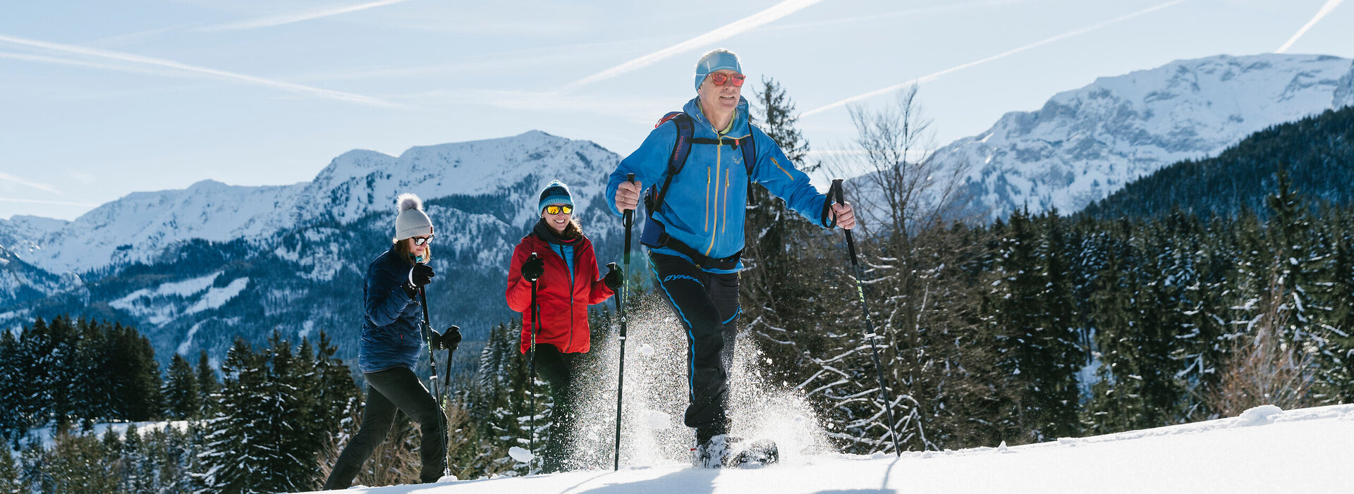 Das Bild zeigt drei Personen in sportlicher Winterkleidung mit Sonnenbrillen und Mützen. Sie laufen hintereinander. Im Hintergrund erheben sich bewaldete, schneebedeckte Berge. Der Himmel ist blau. Die Sonne scheint. Ein Mann läuft voraus. Zwei Frau laufen ihm nach. Alle lächeln. Sie tragen Schneeschuhe und laufen über eine noch unberührte schneebedeckte Wiese. 