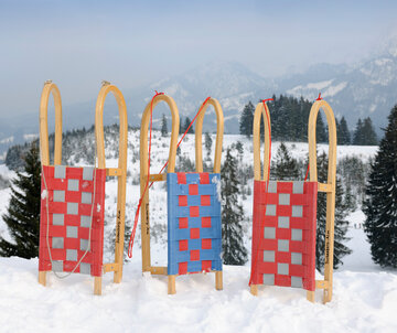 Das Bild zeigt drei Holzschlitten mit bunten Sitzflächen, die an einem Abhang stehen. Rundherum sind hohe Bäume zu sehen. Im Hintergrund erheben sich die schneebedeckten Berge. Die Landschaft ringsum ist tief verschneit. Der Himmel ist blau. Die Sonne scheint. 