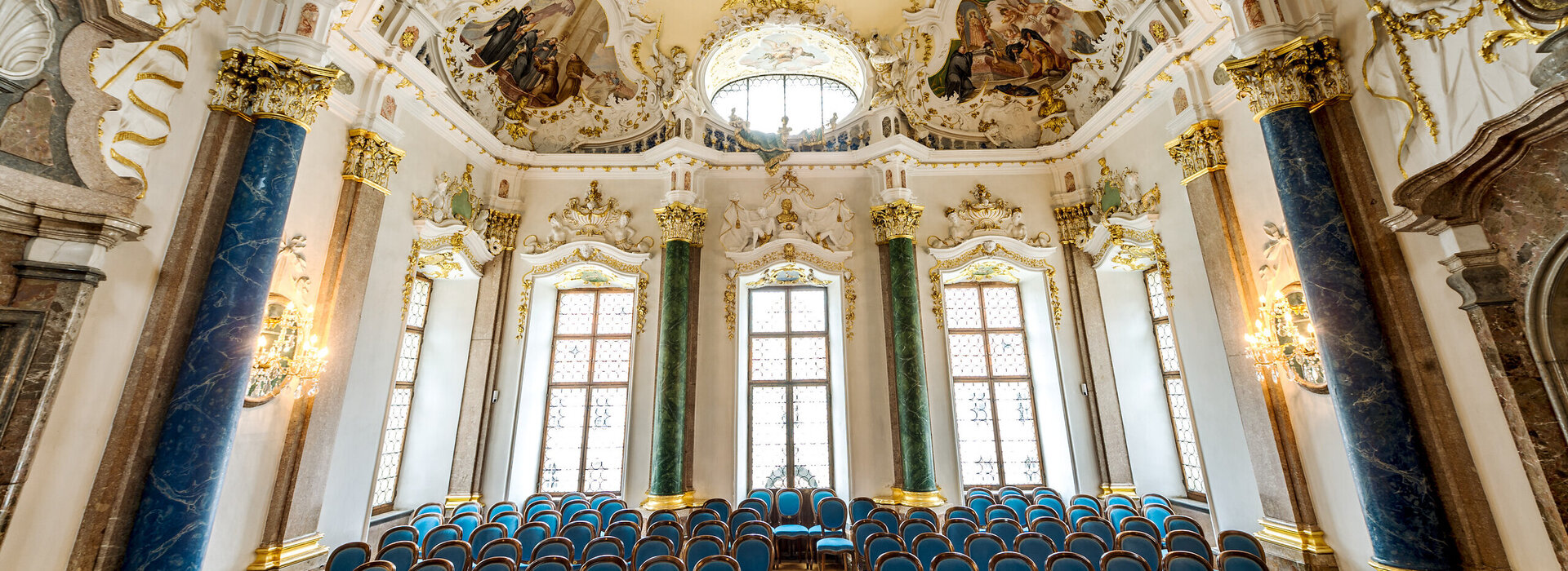 Das Foto zeigt eine prunkvollen Raum. Die Decke ist reich mit Stuck und mehreren Deckengemälden verziert. Den Raum säumen hohe Fenster. Auch die Fensterrahmen sind mit weiß, goldenem Stuck verziert. Der Raum wird von vier Säulen eingerahmt. Zwei Säulen sind Lapislazuli blau, zwei malachitgrün. Der Raum steht voller Holzstühle, die mit blauem Stoff bezogen sind. Die Stühle sind alle Richtung Kamera ausgerichtet. Ein schmaler Gang führt durch die Stühle hindurch. Der Raum ist lichtdurchflutet.  