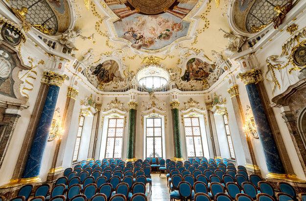Das Foto zeigt eine prunkvollen Raum. Die Decke ist reich mit Stuck und mehreren Deckengemälden verziert. Den Raum säumen hohe Fenster. Auch die Fensterrahmen sind mit weiß, goldenem Stuck verziert. Der Raum wird von vier Säulen eingerahmt. Zwei Säulen sind Lapislazuli blau, zwei malachitgrün. Der Raum steht voller Holzstühle, die mit blauem Stoff bezogen sind. Die Stühle sind alle Richtung Kamera ausgerichtet. Ein schmaler Gang führt durch die Stühle hindurch. Der Raum ist lichtdurchflutet.  