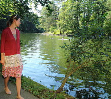 Das Bild zeigt eine Frau auf einem asphaltierten Weg am Ufer eines grünen Sees. Sie ist in einem lebhaften roten Pullover und einem floralen Rock gekleidet. Ihr Blick ist auf den See gerichtet, wo mehrere Personen schwimmen. Der See ist von üppigen grünen Bäumen umgeben und schafft eine ruhige Atmosphäre. 