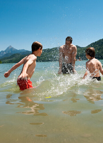 Das Bild zeigt zwei Jungen und einen Mann die im Knietiefen Wasser stehen, Badekleidung tragen und sich gegenseitig nassspritzen. Sie haben sichtlich Spaß. Im Hintergrund sieht man einige Personen schwimmen. Im Hintergrund erheben sich die Berge, davor bewaldete Hügel. Der Himmel ist blau. Die Sonne scheint. 