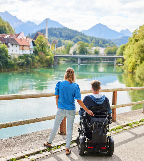 Das Bild fängt einen friedlichen Moment über einer Holzbrücke ein, die einen Blick auf eine Flusslandschaft bietet. Zwei Personen, ein Mann und eine Frau, stehen auf der Brücke, mit dem Rücken zur Kamera. Der Mann sitzt im Rollstuhl, während die Frau neben ihm steht. Beide sind lässig gekleidet, wobei die Frau ein blaues Shirt und der Mann einen schwarzen Mantel trägt. Die Brücke aus Holzdielen spannt sich über den Fluss und bietet einen Aussichtspunkt für den Betrachter. Der Fluss selbst ist ein lebendiges Blau, das den klaren Himmel darüber widerspiegelt. Im Hintergrund schmiegt sich ein malerisches Dorf in die üppige Vegetation, der das Bild mit Charme erfüllt. Die Atmosphäre des Bildes ist eine der Ruhe und Gesellschaft.