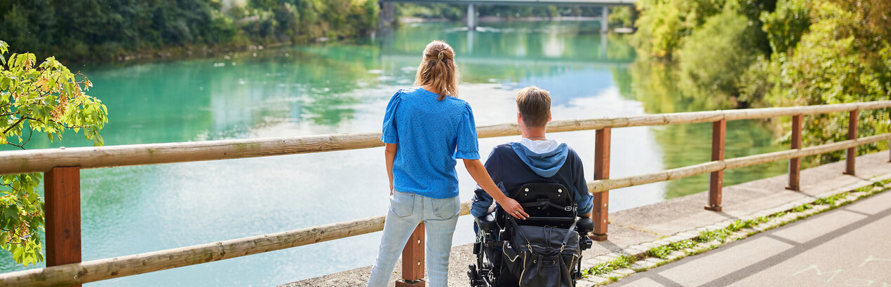 Das Bild fängt einen friedlichen Moment über einer Holzbrücke ein, die einen Blick auf eine Flusslandschaft bietet. Zwei Personen, ein Mann und eine Frau, stehen auf der Brücke, mit dem Rücken zur Kamera. Der Mann sitzt im Rollstuhl, während die Frau neben ihm steht. Beide sind lässig gekleidet, wobei die Frau ein blaues Shirt und der Mann einen schwarzen Mantel trägt. Die Brücke aus Holzdielen spannt sich über den Fluss und bietet einen Aussichtspunkt für den Betrachter. Der Fluss selbst ist ein lebendiges Blau, das den klaren Himmel darüber widerspiegelt. Im Hintergrund schmiegt sich ein malerisches Dorf in die üppige Vegetation, der das Bild mit Charme erfüllt. Die Atmosphäre des Bildes ist eine der Ruhe und Gesellschaft.