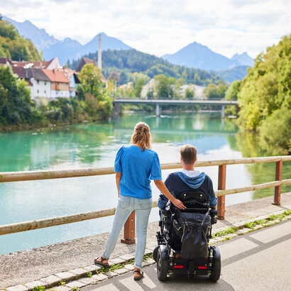 Das Bild fängt einen friedlichen Moment über einer Holzbrücke ein, die einen Blick auf eine Flusslandschaft bietet. Zwei Personen, ein Mann und eine Frau, stehen auf der Brücke, mit dem Rücken zur Kamera. Der Mann sitzt im Rollstuhl, während die Frau neben ihm steht. Beide sind lässig gekleidet, wobei die Frau ein blaues Shirt und der Mann einen schwarzen Mantel trägt. Die Brücke aus Holzdielen spannt sich über den Fluss und bietet einen Aussichtspunkt für den Betrachter. Der Fluss selbst ist ein lebendiges Blau, das den klaren Himmel darüber widerspiegelt. Im Hintergrund schmiegt sich ein malerisches Dorf in die üppige Vegetation, der das Bild mit Charme erfüllt. Die Atmosphäre des Bildes ist eine der Ruhe und Gesellschaft.