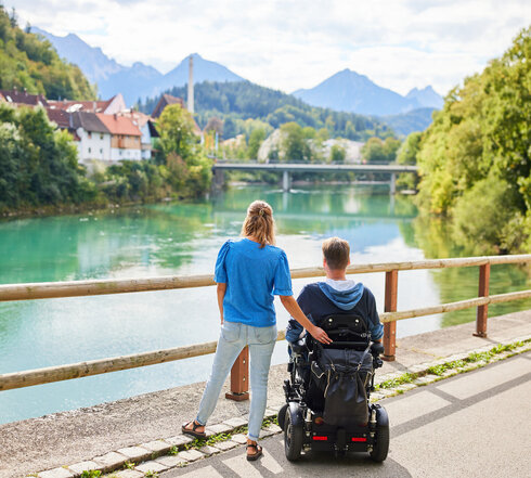 Das Bild fängt einen friedlichen Moment über einer Holzbrücke ein, die einen Blick auf eine Flusslandschaft bietet. Zwei Personen, ein Mann und eine Frau, stehen auf der Brücke, mit dem Rücken zur Kamera. Der Mann sitzt im Rollstuhl, während die Frau neben ihm steht. Beide sind lässig gekleidet, wobei die Frau ein blaues Shirt und der Mann einen schwarzen Mantel trägt. Die Brücke aus Holzdielen spannt sich über den Fluss und bietet einen Aussichtspunkt für den Betrachter. Der Fluss selbst ist ein lebendiges Blau, das den klaren Himmel darüber widerspiegelt. Im Hintergrund schmiegt sich ein malerisches Dorf in die üppige Vegetation, der das Bild mit Charme erfüllt. Die Atmosphäre des Bildes ist eine der Ruhe und Gesellschaft.