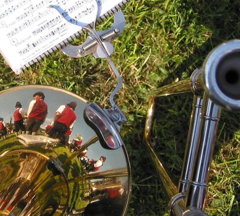 Das Bild zeigt eine Detailaufnahme einer Posaune. Im Trichter der Posaune spiegeln sich in Tracht gekleidete Männer mit Lederhosen, roten Westen, weißen Hemden und Hüten. An der Posaune ist eine Marschgabel befestigt, in der Noten eingespannt sind. Die Posaune liegt auf einer Wiese.  