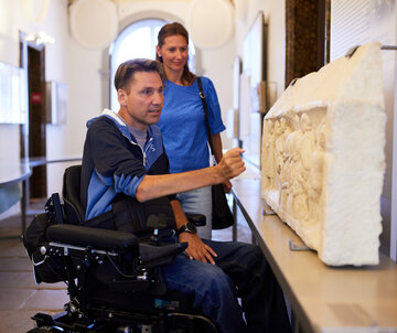 Das Bild erfasst einen Moment in einem Museum, in dem ein Mann im Rollstuhl auf eine große Steinskulptur zeigt. Die Skulptur, die den Mittelpunkt des Bildes darstellt, ist ein großer Steinblock mit komplizierten Schnitzereien. Der Mann, der der Hauptmotiv des Bildes ist, trägt ein blaues Hemd und benutzt seine rechte Hand, um auf die Skulptur zu zeigen. Er sitzt in einem Rollstuhl, der mit einer Rückenlehne und Armlehnen ausgestattet ist.  Im Hintergrund steht eine Frau hinter dem Mann. Sie trägt auch ein blaues Hemd und hält eine schwarze Handtasche. Die Museumsszenerie wird durch die Anwesenheit eines großen Fensters im Hintergrund betont, durch das natürliches Licht die Szene erhellt. Die Gesamtatmosphäre des Bildes ist eine des Neugier und Faszinations, da der Mann und die Frau die komplizierten Details der Skulptur erkunden.