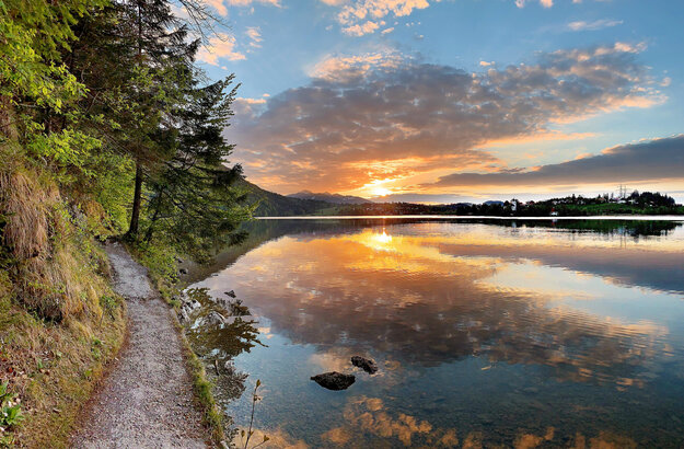 Das Bild zeigt einen See im Abendlicht. Die Sonne geht gerade unter und taucht die Szenerie in ein warmes, fast rötliches Licht. Der Himmel ist blau und mit Wolken durchzogen, die sich durch die Abendsonne orange färben. Am linken Bildrand verläuft ein schmaler, befestigter Uferweg. Das Ufer ist zudem mit Moss, Gräsern und Bäumen bewachsen. Im Hintergrund ist eine kleine Ortschaft zu sehen. Die Wasseroberfläche ist ruhig.  