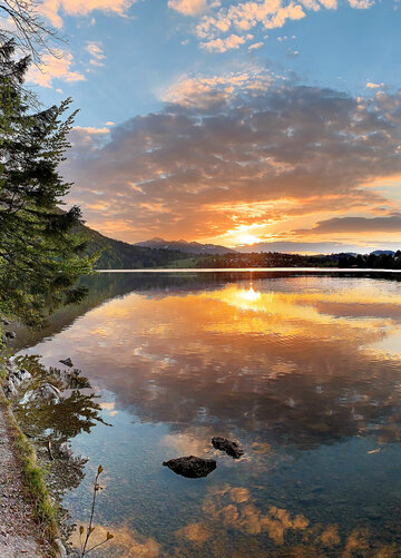 Das Bild zeigt einen See im Abendlicht. Die Sonne geht gerade unter und taucht die Szenerie in ein warmes, fast rötliches Licht. Der Himmel ist blau und mit Wolken durchzogen, die sich durch die Abendsonne orange färben. Am linken Bildrand verläuft ein schmaler, befestigter Uferweg. Das Ufer ist zudem mit Moss, Gräsern und Bäumen bewachsen. Im Hintergrund ist eine kleine Ortschaft zu sehen. Die Wasseroberfläche ist ruhig.  