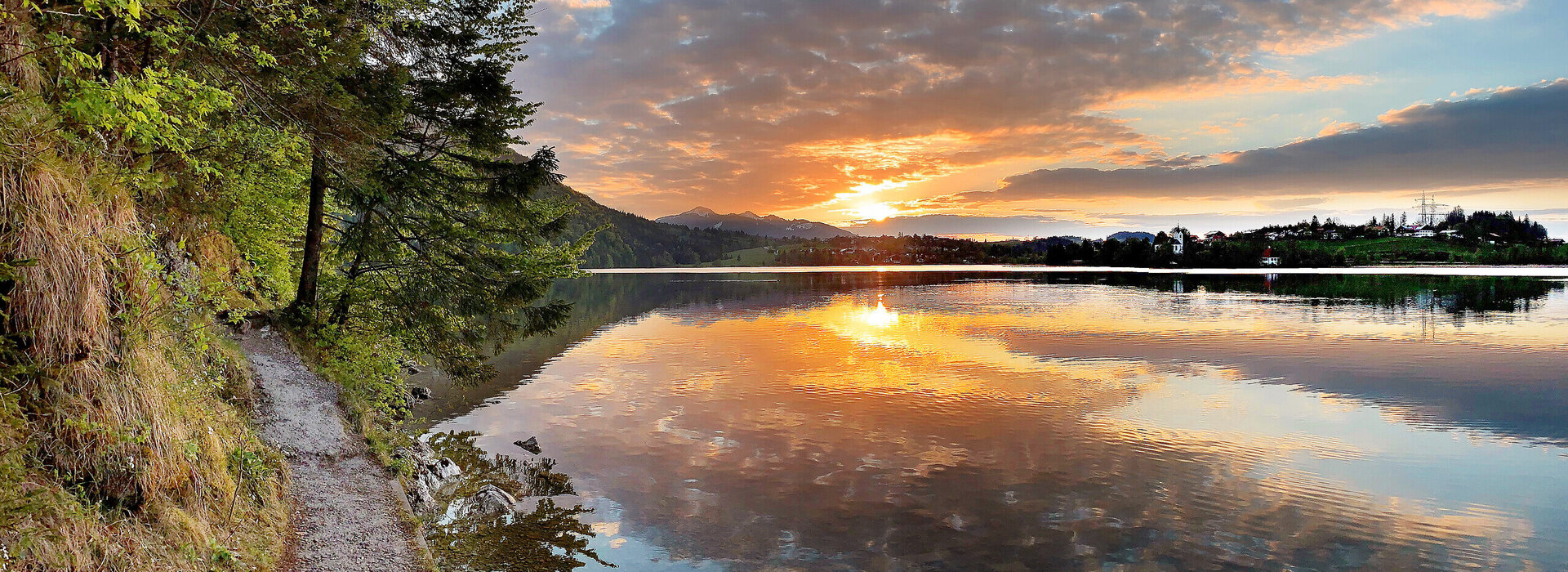 Das Bild zeigt einen See im Abendlicht. Die Sonne geht gerade unter und taucht die Szenerie in ein warmes, fast rötliches Licht. Der Himmel ist blau und mit Wolken durchzogen, die sich durch die Abendsonne orange färben. Am linken Bildrand verläuft ein schmaler, befestigter Uferweg. Das Ufer ist zudem mit Moss, Gräsern und Bäumen bewachsen. Im Hintergrund ist eine kleine Ortschaft zu sehen. Die Wasseroberfläche ist ruhig.  