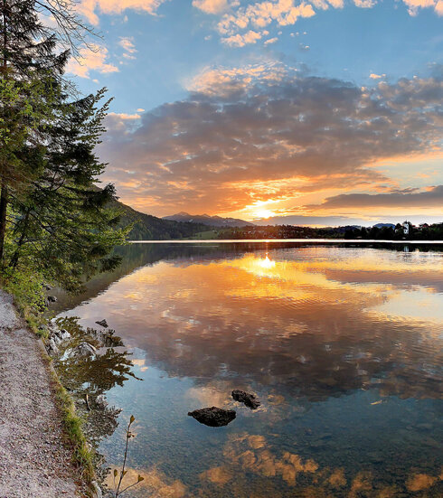 Das Bild zeigt einen See im Abendlicht. Die Sonne geht gerade unter und taucht die Szenerie in ein warmes, fast rötliches Licht. Der Himmel ist blau und mit Wolken durchzogen, die sich durch die Abendsonne orange färben. Am linken Bildrand verläuft ein schmaler, befestigter Uferweg. Das Ufer ist zudem mit Moss, Gräsern und Bäumen bewachsen. Im Hintergrund ist eine kleine Ortschaft zu sehen. Die Wasseroberfläche ist ruhig.  