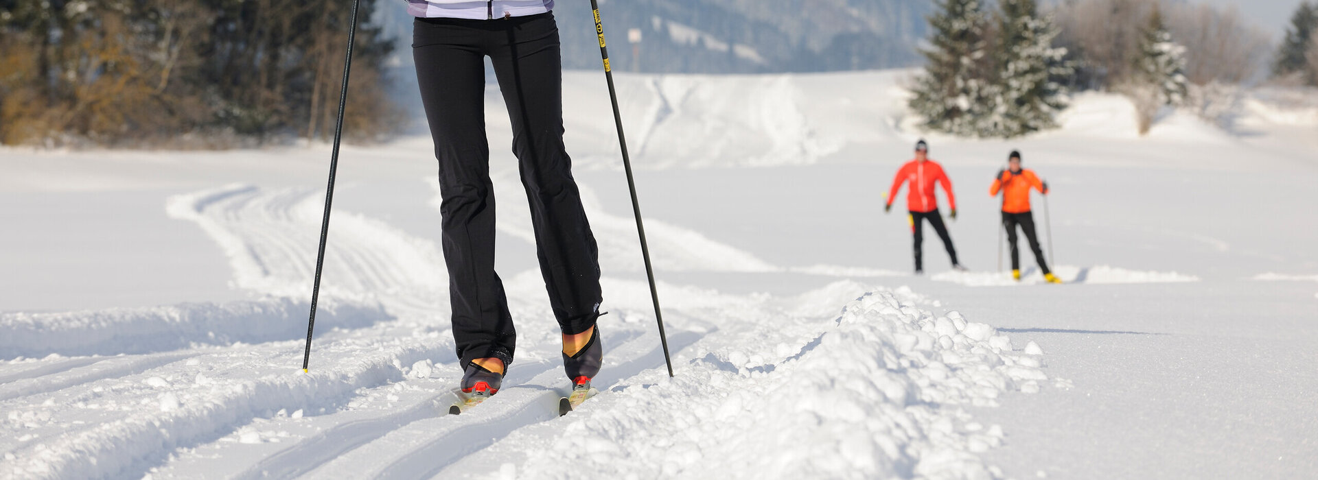 Das Bild zeigt drei Personen beim Ski-Langlauf auf einer Langlaufloipe. Im Vordergrund fährt eine Frau. Im Hintergrund sind weitere zwei Personen zu sehen.  Die Landschaft ringsum ist tief verschneit. Im Hintergrund erheben sich die schneebedeckten Berge. Der Himmel ist blau. Die Sonne scheint.
