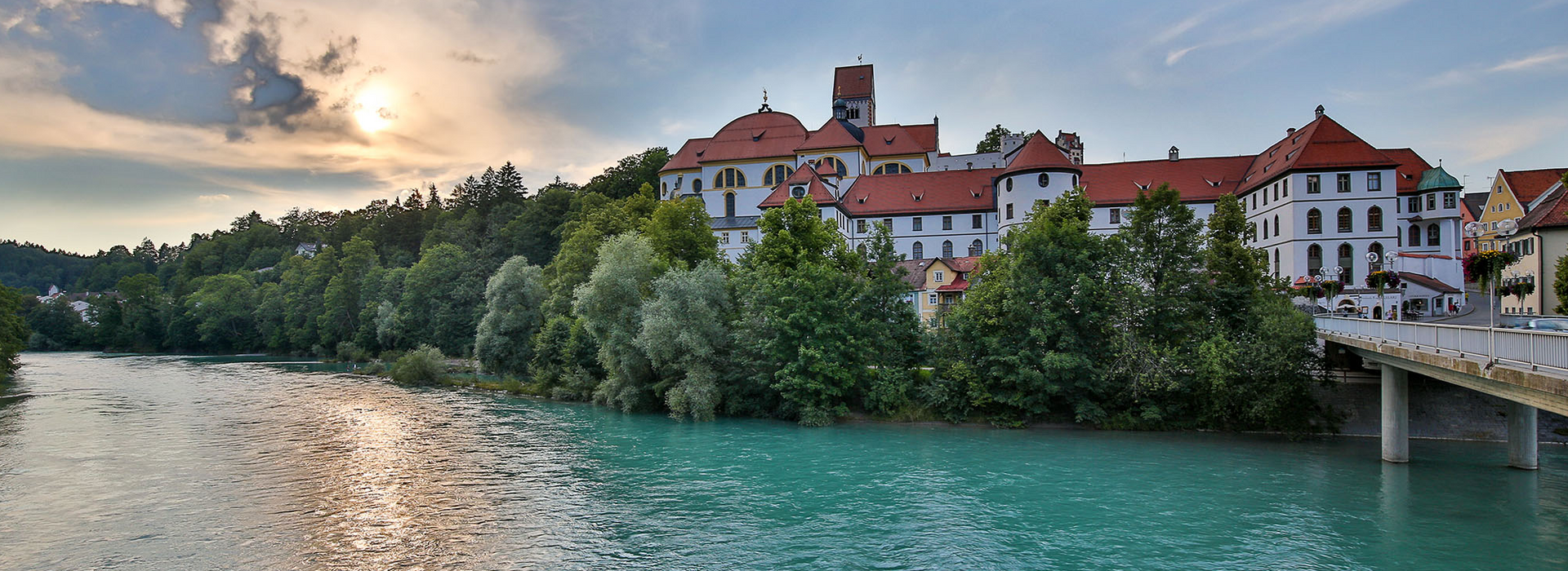 Das Bild zeigt ein schlossähnliches Gebäude, das auf einer Anhöhe am Ufer des Flusses Lech thront. Bei dem Gebäudekomplex handelt es sich um das ehemalige Benediktinerkloster St. Mang, das mit seinem roten Dach, majestätisch vor dem Hintergrund eines klaren blauen Himmels steht. Der türkise Fluss, fließt sanft vor dem Gebäude und spiegelt das Sonnenlicht. Die umgebende Landschaft ist üppig grün, mit Bäumen und Büschen, die die Landschaft prägen. Die untergehende Sonne tauch die Szene in warmes Licht.