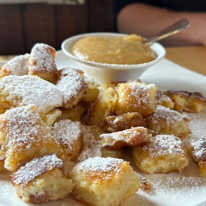 Das Foto zeigt die Mehlspeise Kaiserschmarrn. Die mit weißem Puderzucker überzogen ist. Die Speise ist auf einem weißen Teller angerichtet. Ebenso auf dem Teller steht ein weißes Schälchen mit Apfelmus. 