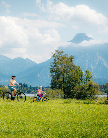 Das Bild fängt eine friedvolle Szene in einer grünen Wiese ein, wo drei Personen ein Fahrrad ausführen. Die Wiese ist üppig und grün, sie bietet eine lebendige Kulisse zur Szene. Der Himmel über ist ein klares Blau, garniert mit flauschigen weißen Wolken. In der Ferne erheben sich majestätische Berge gegen den Himmel, was der Szene ein Gefühl der Größe verleiht. Die Personen im Bild sind in lässiger Kleidung gekleidet, was auf einen gemütlichen Tag im Freien hinweist. Die Fahrräder, auf denen sie fahren, sind gut sichtbar, wobei eine Person ein Fahrrad reitet und die anderen beiden zu Fuß gehen. Die allgemeine Atmosphäre des Bildes ist die einer Ruhe und des Genusses der Natur.