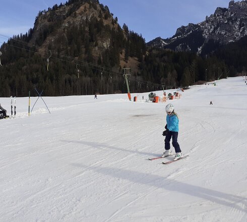 Das Bild fängt eine lebendige Winterszene auf einem verschneiten Berg ein. In der Mitte des Bildes ist ein junges Mädchen dabei, den Hang mit Skiern hinunter zu fahren. Sie ist in einem blauen Jackett und einem weißen Helm gekleidet und trägt Skistöcke. Ihre Skier sind klar sichtbar und schneiden durch den unberührten weißen Schnee.  Im Hintergrund sind mehrere andere Skifahrer zu sehen, die den Berg genießen. Sie sind über den Hang verstreut, wobei einige näher an der Kamera und andere weiter weg sind. Der Berg selbst ist dick mit Schnee bedeckt und durchsetzt mit Bäumen, die eine grüne Nuance zur ansonsten weißen Landschaft hinzufügen.  Der Himmel über ist ein klarer blauer Ton, was auf einen hellen und sonnigen Tag hindeutet. Die Gesamtszene bietet ein Bild von Aufregung und Freude, da die Menschen das schöne Winterwetter nutzen, um sich im Freien zu betätigen.