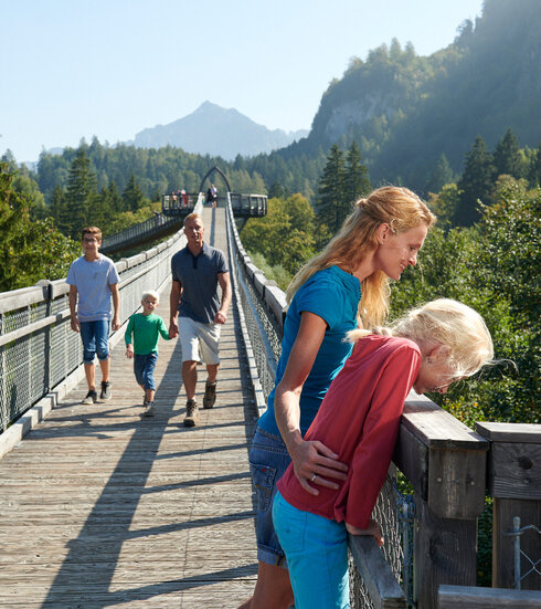 Das Bild fängt eine friedliche Szene auf einer Holzbrücke ein. Ein Familienvater ist mit einer blauen Jacke als dominantes Motiv abgebildet und führt die Familie über die Brücke. Seine Frau mit einer rosafarbenen Jacke folgt ihm. Ihre beiden Kinder, eine in einer grünen Jacke und die andere in einer roten Jacke, laufen dicht hinter ihnen. Die Brücke, die sie betreten haben, ist aus Holz und wird von üppig grünen Bäumen und einem klaren blauen Himmel umgeben. In der Ferne sind majestätische Berge zu sehen, die zur Schönheit der Szene beitragen. Die Familie scheint die Zeit miteinander zu genießen und erfreut sich am beeindruckenden Blick auf die Berge.