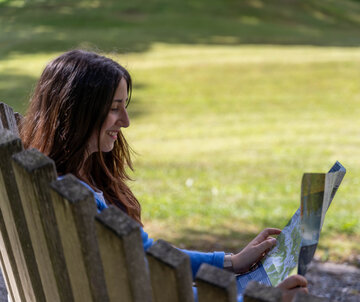 Das Bild zeigt eine Frau, die einen Flyer liest. Man sieht die Frau nur von hinten. Sie sitzt auf einer Holzbank und lächelt. Im Hintergrund ist eine Wiese zu sehen. Die Sonne scheint.  