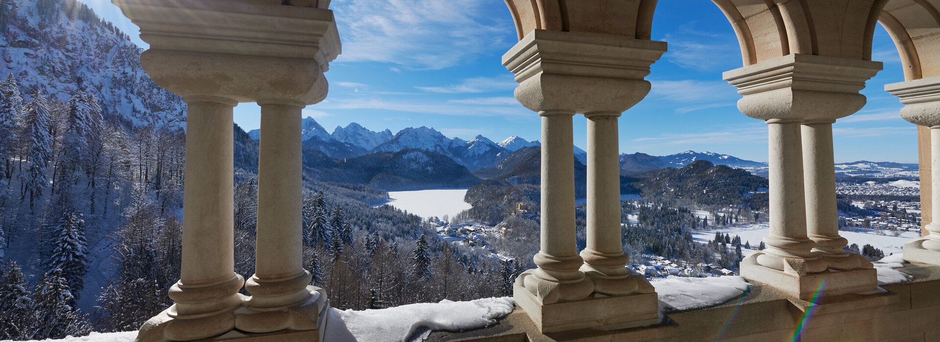 Das Bild fängt einen atemberaubenden Blick auf eine schneebedeckte Bergkette ein, die vom hohen Aussichtspunkt aus betrachtet wird. Die Perspektive ist von einem Balkon aus, der mit einer Reihe von Bögen geschmückt ist. Diese Bögen, aus Stein gefertigt, sind gleichmäßig gesetzt und verleihen der Szene ein Gefühl von Symmetrie und Gleichgewicht. Die Sonne scheint hell, wirft ein warmes Glühen auf die schneebedeckten Berge und beleuchtet die Bögen so, dass ihre architektonische Schönheit betont wird.