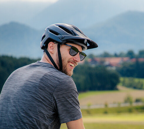 Das Bild zeigt einen Mann in Freizeitkleidung und Fahrradhelm beim Radfahren. Er blickt über seine rechte Schulter zum Betrachter und lacht. Er fährt auf einer befestigen Straße. Links und rechts neben der Straße grenzen Wiesen an. Im Hintergrunderheben sich Berge und bewaldete Hügel. Die Sonne scheint. 