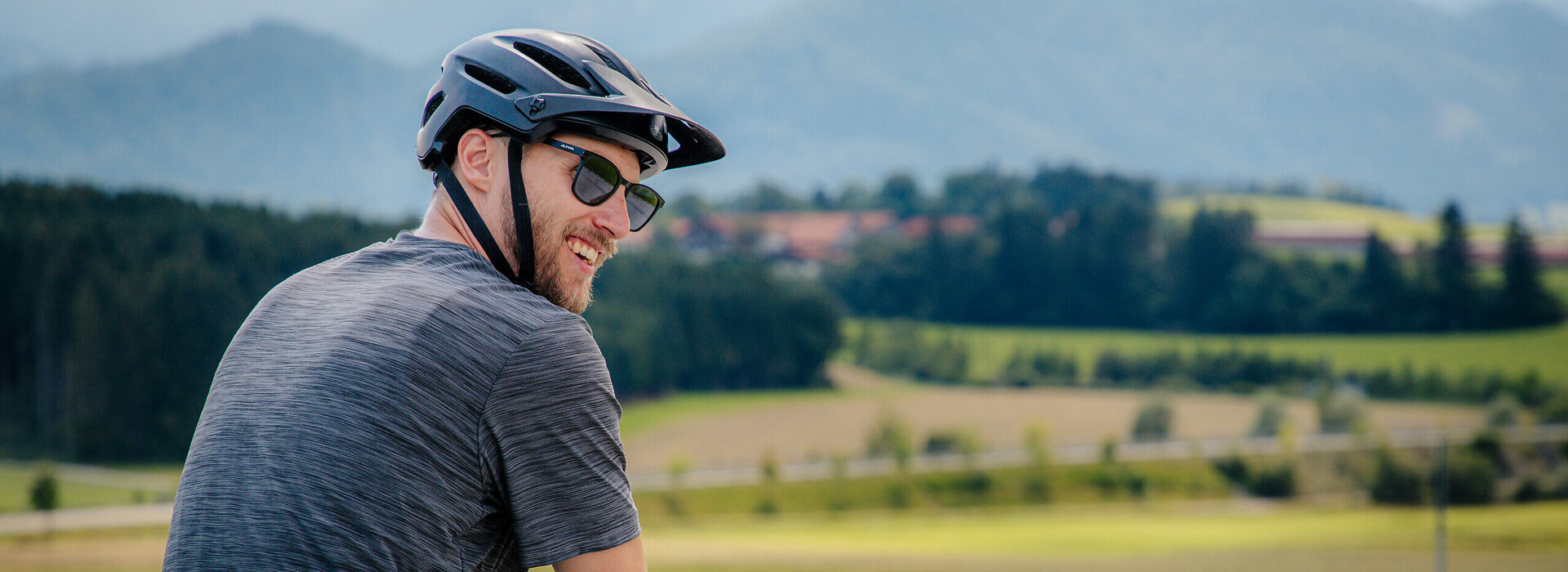 Das Bild zeigt einen Mann in Freizeitkleidung und Fahrradhelm beim Radfahren. Er blickt über seine rechte Schulter zum Betrachter und lacht. Er fährt auf einer befestigen Straße. Links und rechts neben der Straße grenzen Wiesen an. Im Hintergrunderheben sich Berge und bewaldete Hügel. Die Sonne scheint. 