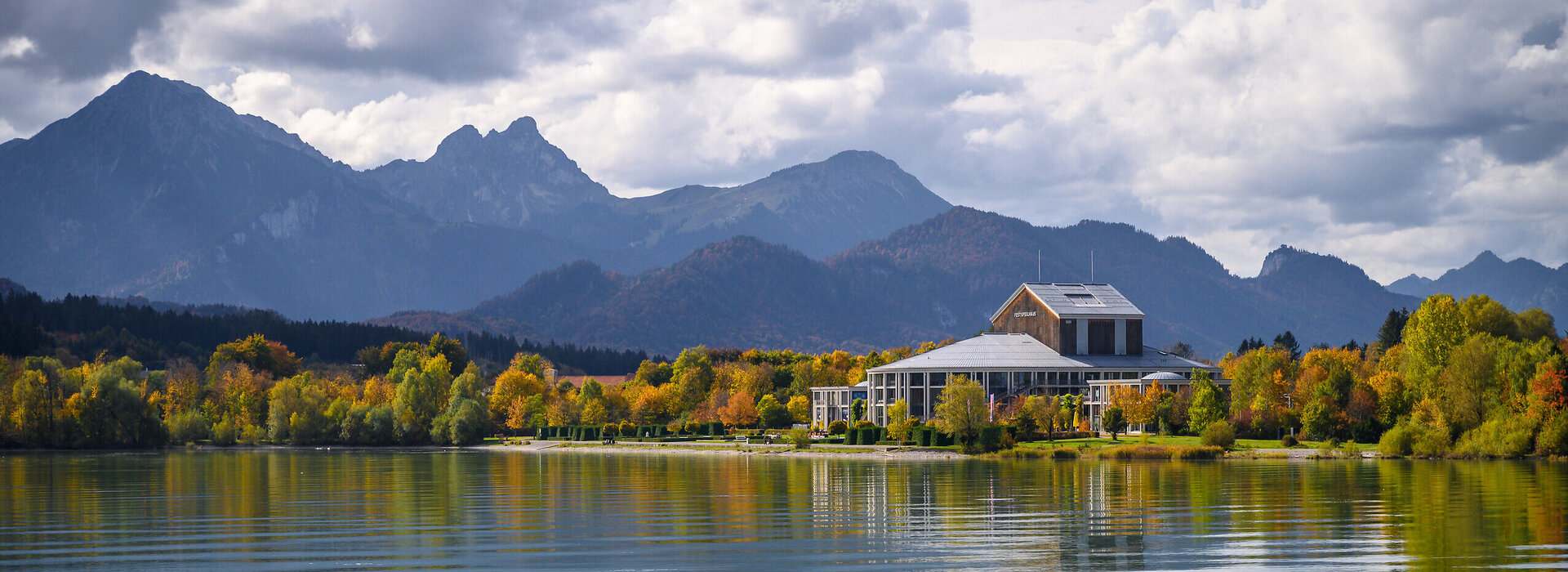 Das Foto wurde vom See aus aufgenommen. Am Ufer steht ein prächtiges Gebäude mit einem grauen Dach und vielen Fenstern. Der Mittelteil des Gebäudes ist rund. Es handelt sich um das Festspielhaus Neuschwanstein. Die Bäume ringsum haben schon eine bunte Farbe angenommen. Im Hintergrund erstreckt sich die Alpenkette. Der Himmel ist wolkenverhangen, trotzdem scheint die Sonne. 