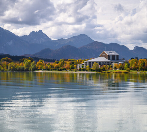 Das Foto wurde vom See aus aufgenommen. Am Ufer steht ein prächtiges Gebäude mit einem grauen Dach und vielen Fenstern. Der Mittelteil des Gebäudes ist rund. Es handelt sich um das Festspielhaus Neuschwanstein. Die Bäume ringsum haben schon eine bunte Farbe angenommen. Im Hintergrund erstreckt sich die Alpenkette. Der Himmel ist wolkenverhangen, trotzdem scheint die Sonne. 