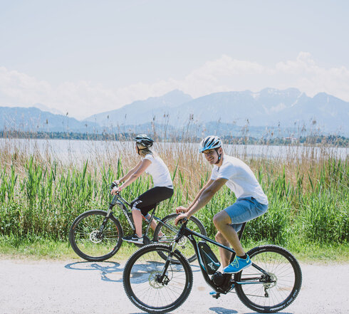Das Bild zeigt eine zwei E-Bikefahrer. Einen Mann und eine Frau in kurzer Freizeitkleidung. Beide Personen tragen einen Helm. Der Mann blickt zum Betrachter. Sie fahren auf einer unbefestigten Straße, an die parallel ein Schilfgürtel anschließt. Im Anschluss an den Schilfgürtel ist ein See zu sehen. Im Hintergrund erheben sich Berge und bewaldete Hügel. Die Sonne scheint. Der Himmel ist blau.  