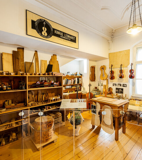 Man blickt in eine Geigenbauerwerkstatt. Viele Geigen und Bratschen hängen an einer Schnur nebeneinander. Ein Regal ist mit Hölzern, Schablonen und Latten gefüllt. Ein Regal mit Gläsern und Flaschen voller Flüssigkeiten. Vor einem Fenster steht eine Werkbank. Über der Szenerie hängt ein Firmenschild. Darauf steht: „Konrad Leonhart, Geigenbaumeister.“ Am Schreibtisch im Hintergrund lehnt ein Cello. Das Regal über dem Schreibtisch ist mit Büchern und Ordnern gefüllt. 