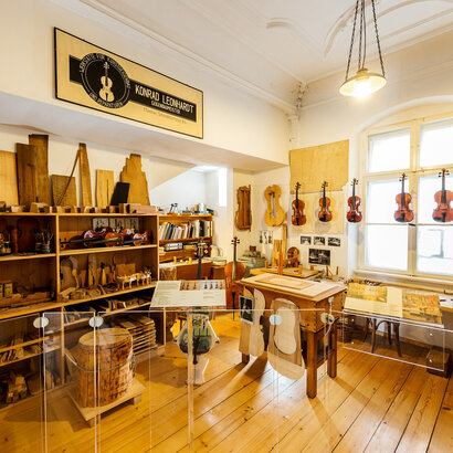 Man blickt in eine Geigenbauerwerkstatt. Viele Geigen und Bratschen hängen an einer Schnur nebeneinander. Ein Regal ist mit Hölzern, Schablonen und Latten gefüllt. Ein Regal mit Gläsern und Flaschen voller Flüssigkeiten. Vor einem Fenster steht eine Werkbank. Über der Szenerie hängt ein Firmenschild. Darauf steht: „Konrad Leonhart, Geigenbaumeister.“ Am Schreibtisch im Hintergrund lehnt ein Cello. Das Regal über dem Schreibtisch ist mit Büchern und Ordnern gefüllt. 