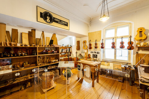 Man blickt in eine Geigenbauerwerkstatt. Viele Geigen und Bratschen hängen an einer Schnur nebeneinander. Ein Regal ist mit Hölzern, Schablonen und Latten gefüllt. Ein Regal mit Gläsern und Flaschen voller Flüssigkeiten. Vor einem Fenster steht eine Werkbank. Über der Szenerie hängt ein Firmenschild. Darauf steht: „Konrad Leonhart, Geigenbaumeister.“ Am Schreibtisch im Hintergrund lehnt ein Cello. Das Regal über dem Schreibtisch ist mit Büchern und Ordnern gefüllt. 