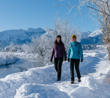 Das Bild zeigt eine friedliche Winterlandschaft. Zwei Frauen in Winterklamotten sind auf einem mit Schnee bedeckten Pfad zu sehen. Die Person auf der linken Seite trägt eine lilafarbene Winterjacke, während diejenige auf der rechten Seite eine blaue Winterjacke trägt. Der Weg, den sie entlanglaufen, ist mit Schnee bedeckt, nur ihre Fußabdrücke sind sichtbar. Der Hintergrund des Bildes ist ein atemberaubender Blick auf schneebedeckte Berge, was zur Ruhe der Szene beiträgt. Der Himmel ist klar und blau, frei von Wolken. Das Gesamtbild zeichnet ein Bild eines friedlichen Wintertages, mit den beiden Personen, die ihren Spaziergang inmitten der Schönheit der Natur genießen.